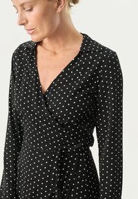 Schwarzes Wickelkleid mit langen Ärmeln, das weiße Polka-Dots, einen Kragen und einen Bindegürtel in der Taille für eine figurbetonte Silhouette aufweist.