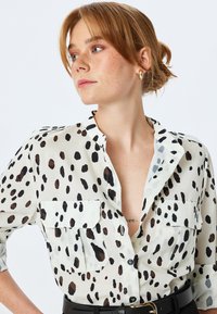 Lättvikts, genomskinlig blus i krämfärg med svarta och bruna oregelbundna fläckar, knäppning fram, två bröstfickor och en avslappnad krage.