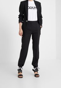 Blazer negro ajustado sobre una camiseta blanca con el texto "HUGO", combinado con pantalones negros que presentan acentos en blanco y negro en los puños.