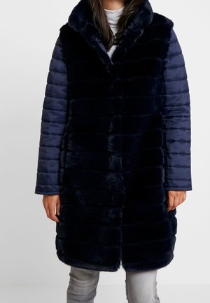 Femme portant un long manteau en fausse fourrure bleu marine foncé avec des manches matelassées rembourrées et un col montant, par-dessus un haut blanc et un jean gris.