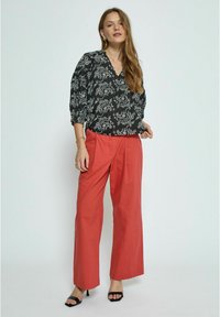 Schwarze Bluse mit weißem Paisleymuster, V-Ausschnitt und Puffärmeln; kombiniert mit hochgafften, weit geschnittenen korallenfarbenen Hosen und schwarzen Absatzsandalen.