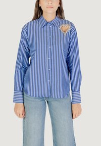 Camicia a maniche lunghe a righe blu e bianche con apertura a bottoni, caratterizzata da un'applicazione a forma di cuore sul petto e maniche con polsini.