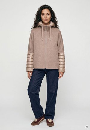 Frau mit dunklem lockigem Haar, die eine beiger Reißverschlussjacke mit Steppärmeln, dunkelblaue Jeans und braune Sneakers trägt, steht vor weißem Hintergrund.