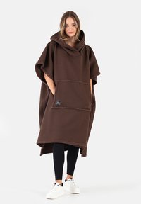 Brązowy polarowy poncho z kapturem, dużą przednią kieszenią i luźnym krojem. Posiada szerokie rękawy oraz miękką teksturę. Noszone na czarnych legginsach.