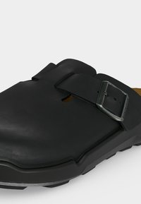 Μαύρο δερμάτινο κλογκ με ρυθμιζόμενο λουράκι με αγκράφα και υφή από καουτσούκ στη σόλα για καλύτερη πρόσφυση σε λευκό φόντο.