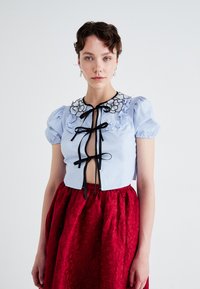 Light blue cropped blouse with puff sleeves, adorned with black floral embroidery and bows, paired with a high-waisted red skirt featuring floral patterns.