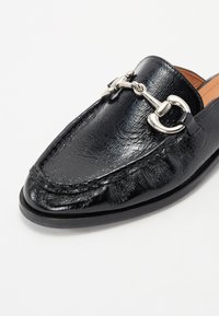 Black leather loafer with a shiny finish, featuring decorative silver hardware and a textured design across the upper. Low-profile, sleek shape.