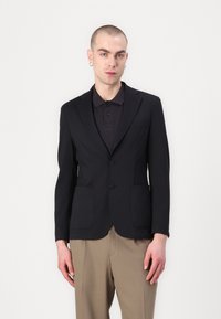 Must mustriga bleiser koos kahe esikotiga, sälgustatud kraega ja kahe nööbiga. Kavam dressitud musta polo särgi ja beežide pükstega.