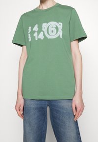 Groene katoenen T-shirt met een gedrukt ontwerp van witte cijfers, met korte mouwen en een ronde hals. Gekleed met blauwe denim jeans.