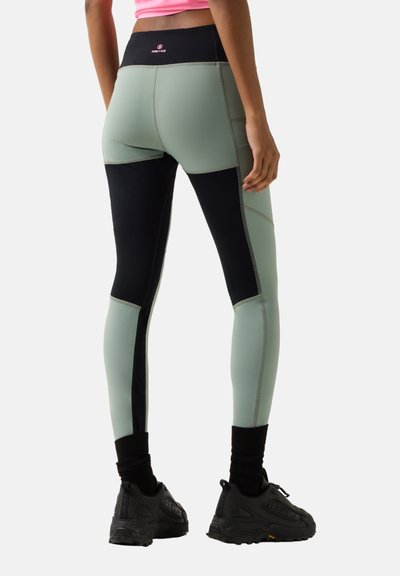 Kvinde iført tofarvede tætsiddende leggings i grøn og sort, sorte sneakers og en lyserød crop top, stående med ryggen vendt mod kameraet.