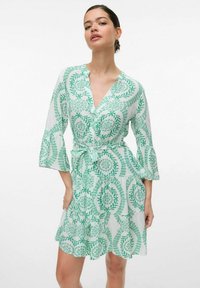 Grön och vit mönstrad klänning med trekvarts ärmar, V-ringning och knytbälte i midjan. Tyget verkar lätt och flödande.