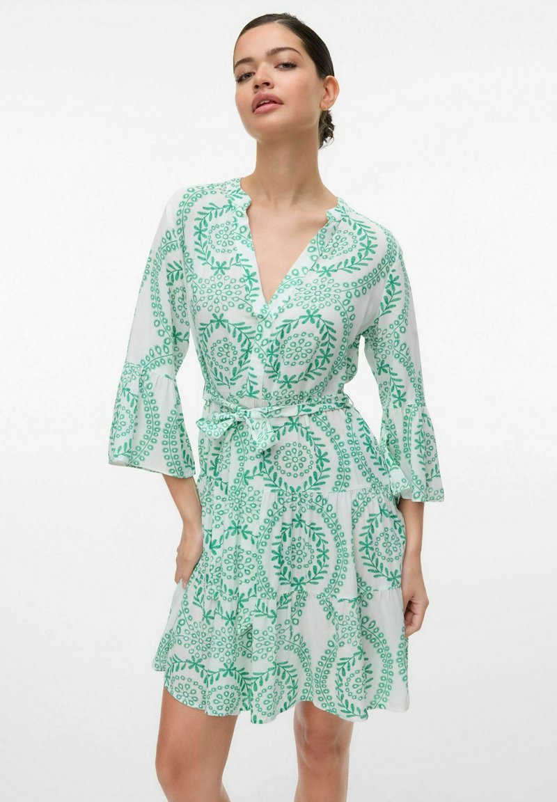 Grön och vit mönstrad klänning med trekvarts ärmar, V-ringning och knytbälte i midjan. Tyget verkar lätt och flödande.