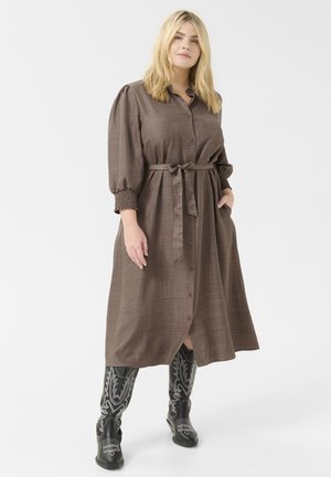 Lång brun klänning med knappframsidan, knytmidja och puffärmar; har sidofickor. Modellen bär svarta broderade cowboystövlar.