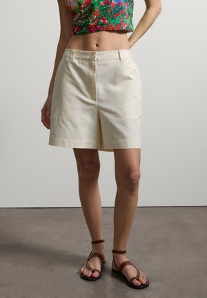 Frau trägt beige High-Waist-Shorts, florales Crop-Top und braune flache Sandalen mit Riemchen, steht auf grauem Boden vor einer schlichten Wand.