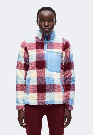 Personne portant une veste polaire à carreaux multicolores avec une poche poitrine zippée bleu clair et un pantalon bordeaux, debout devant un fond uni.
