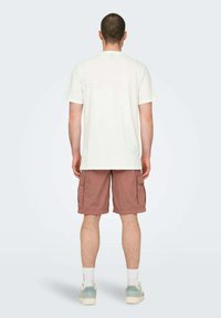 Camiseta blanca de algodón con mangas cortas, combinada con pantalones cortos cargo de color rosa claro. El modelo lleva calcetines blancos y zapatos de color azul claro.