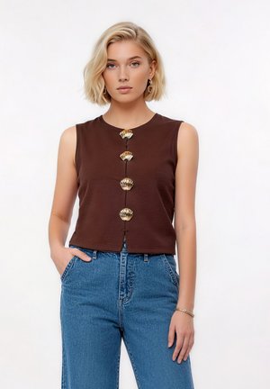 Mujer rubia con top marrón sin mangas con botones dorados en forma de concha y vaqueros azules de talle alto, de pie frente a un fondo liso.