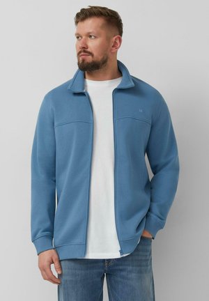 Uomo che indossa una giacca con zip azzurro chiaro sopra una maglietta bianca e jeans blu, in piedi con una mano nella tasca, guarda verso sinistra.