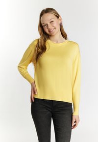Keltainen neulepaita, jossa on pitkät hihat ja pyöreä kaula-aukko. Se on rento malli ja siinä on hillitty ribbaus helman kohdalla. Yhdistetty mustiin farkkuihin.