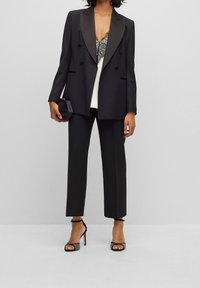 Černé upravené sako s lesklými saténovými klopami, doplněné úzkými černými kalhotami a vzorovaným krajkovým topem. Modelka drží malou černou kabelku.