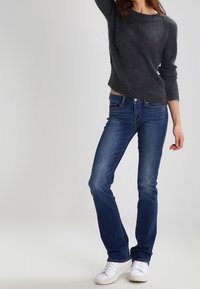 Pull gris en maille avec ourlet côtelé, associé à un jean droit bleu et des baskets blanches. Le pull a des manches longues et une coupe décontractée.
