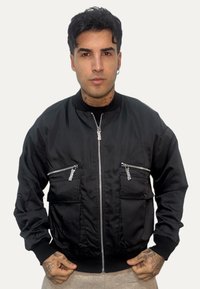 Giacca bomber nera in tessuto liscio, con due tasche sul petto con cerniera, polsini e collo a costine, chiusura frontale con zip.