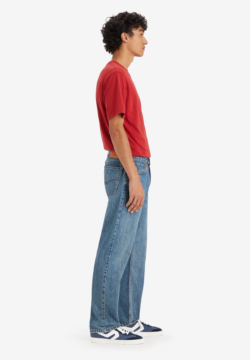 Camiseta de algodón roja, corte holgado, combinada con jeans rectos de mezclilla azul claro. Zapatillas blancas con detalles en azul marino en la suela.