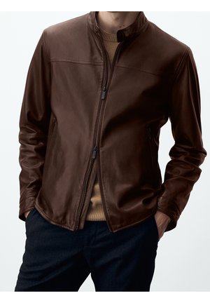 Homme portant une veste en cuir marron foncé zippée par-dessus un pull en maille beige et un pantalon foncé, les mains dans les poches de la veste.