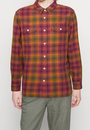 Mann trägt ein langärmeliges Hemd mit kariertem Muster in Rot, Orange, Grün und Gelb sowie graue Hose und steht vor einer schlichten Wand.