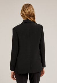 Blazer negro ajustado con solapa muesca, hombros estructurados y una textura suave. Presenta una única abertura trasera y mangas largas.