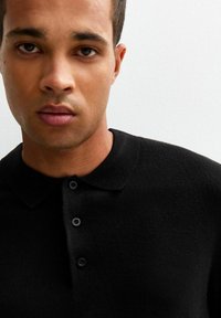 Giovane uomo con carnagione media che indossa una camicia nera con colletto e bottoni, rivolto in avanti contro uno sfondo bianco semplice.