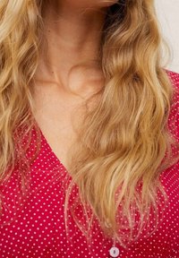 Czerwona bluzka w kropki z dekoltem w serek, z lekkiego materiału o subtelnej teksturze. Faliste blond włosy częściowo widoczne wokół dekoltu.