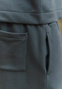 Tejido de sudadera gris oscuro con un bolsillo lateral y un detalle de marca. Textura suave con un ajuste relajado y un diseño de costuras.