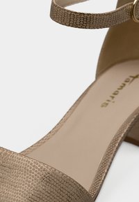 Μπεζ ψηλοτάκουνο σανδάλι με υφή που μοιάζει με δέρμα ερπετού, ρυθμιζόμενο λουράκι στον αστράγαλο και λείο εσωτερικό πέλμα που παρουσιάζει ένα χρυσό λογότυπο.