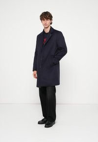 Marineblauer Mantel aus Wollmischung mit einreihigem Design, spitzem Revers und Seitentaschen. Kombiniert mit schwarzer Hose und Schuhen.