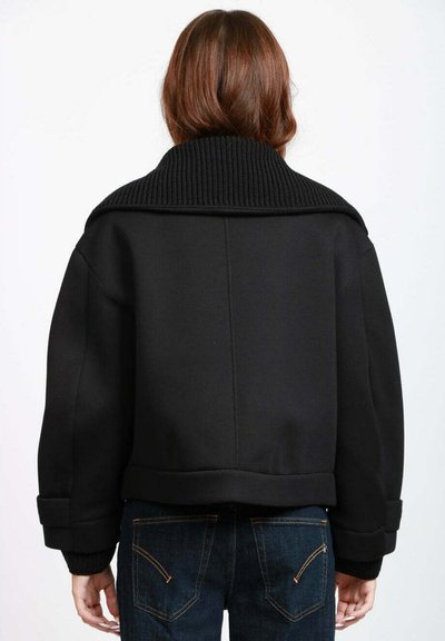 Giacca nera corta con colletto a coste, vestibilità oversize e texture liscia. Abbinata a jeans di denim blu scuro con dettagli cuciti.
