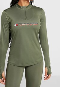 Olivgrünes, sportliches Langarmshirt mit halbem Reißverschluss am Kragen und "TOMMY SPORT"-Logo auf der Brust. Texturiertes, elastisches Material.
