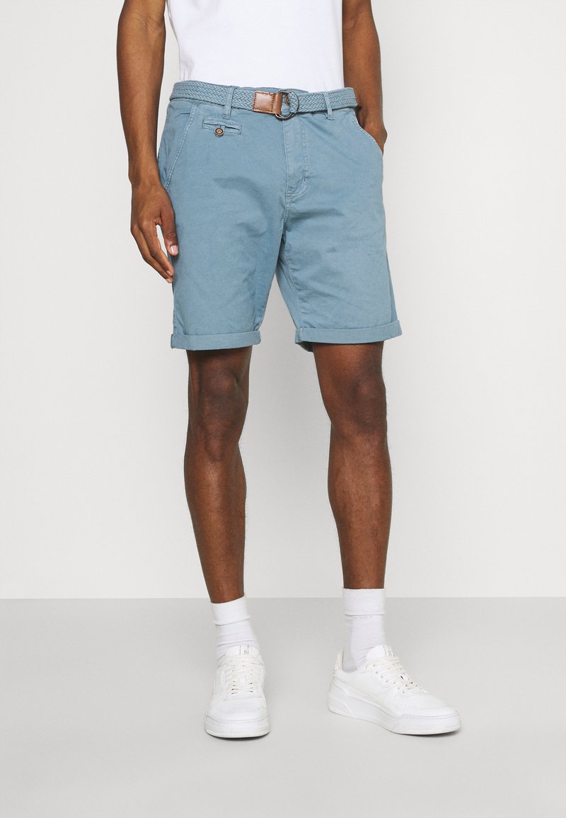 Pantalones cortos de color azul claro hechos de algodón, con un cinturón trenzado que tiene un acento marrón. Los puños enrollados realzan el diseño casual. Se combinan con zapatillas blancas.