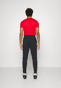 T-shirt sportif rouge ajusté à manches courtes, associé à un pantalon de jogging noir fuselé. Modèle portant des chaussures de sport noires avec des accents blancs et orange.