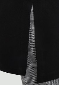 Svart plagg med en sidoklyfta som avslöjar mönstrad grå och vit rutigt tyg under. Slät yta, skräddarsydd design och rena linjer.