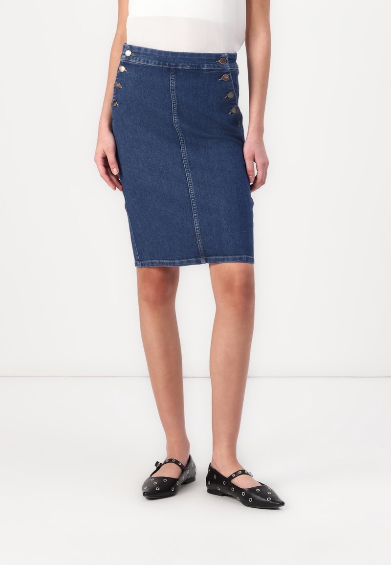 Femme portant une jupe en denim bleu longueur genoux avec six boutons sur les côtés devant, un haut blanc et des ballerines noires avec des détails œillets.