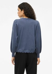 Mujer con cabello rizado, vestida con un suéter de mangas largas azul con un patrón y pantalones negros, de espaldas contra un fondo blanco.