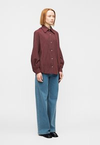 Chemise boutonnée avec des rayures verticales bordeaux et blanches, manches longues bouffantes et col pointu. Associée à un jean bleu taille haute à jambe large.