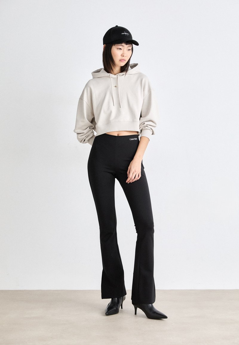 Beige Kapuzenpullover mit Kordelzug, schwarze hochtaillierte, ausgestellte Leggings und schwarze spitze Absätze. Einfaches Design, keine sichtbaren Muster oder Logos.