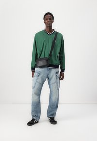 Cămașă verde cu mânecă lungă și guler alb-negru, blugi cargo largi de culoare albastru deschis, geantă crossbody neagră și adidași negri.