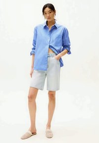 Femme debout dans une chemise à rayures bleues, un short en denim clair et des chaussures slip-on tressées beiges, devant un fond blanc uni.