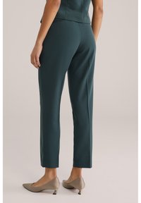 Vrouw draagt donkergroene rechte broekspijpen en beige kittenhakken, staand tegen een effen lichte achtergrond.