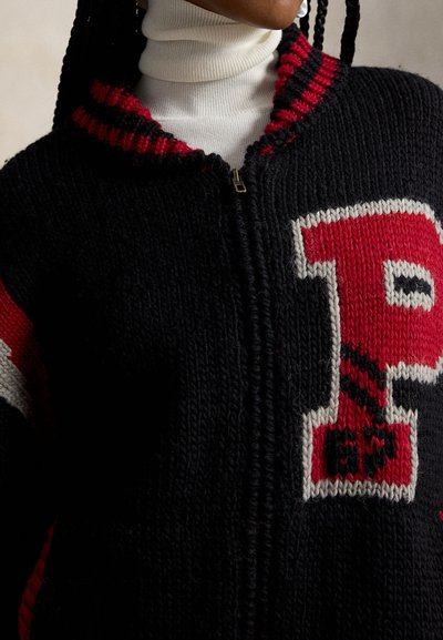 Κοντινό πλάνο ενός μαύρου πλεκτού φούτερ με φερμουάρ και κόκκινο-λευκή μπαλώματος με το γράμμα «P», που φοριέται πάνω από έναν λευκό ζιβάγκο, με ορατά πλεγμένα μαλλιά.