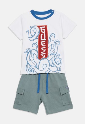 Weißes Kurzarmhemd mit blauen Oktopus-Tentakeln und rotem vertikalem "GUESS"-Logo, kombiniert mit grünen Shorts mit seitlichen Taschen und blauen Kordeln.