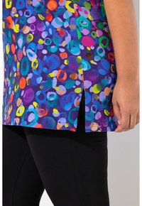 Farverigt top med en lilla baggrund og cirkulære mønstre i rød, blå, grøn og gul. Sideåbning detalje, bæres over sorte leggings.